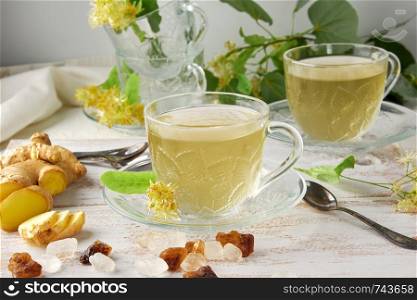 transparent cup with tea from ginger and linden on a white wooden board, next to pieces of sugar and twigs with green leaves