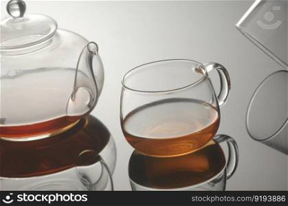Transparent glass teapot and cup of tea on the reflective surface on a light gray background. Transparent glass teapot and cup with tea
