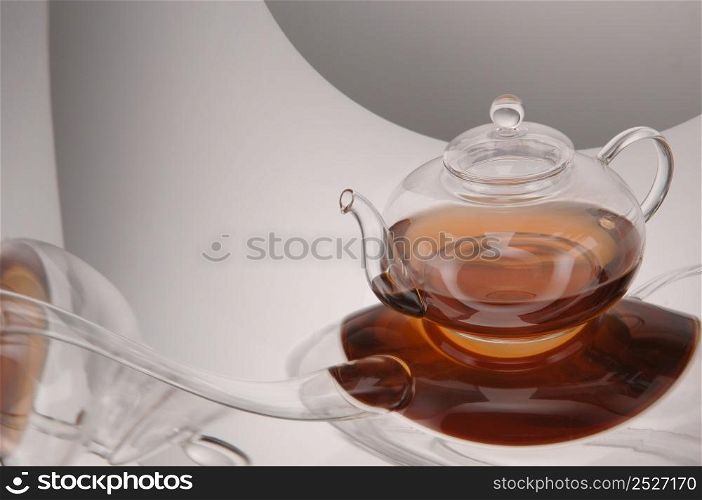 Transparent glass teapot with tea on the reflective surface on a light gray background. Transparent glass teapot and cup with tea