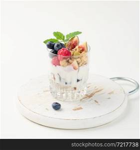 transparent glass with granola poured with yogurt, on top of ripe raspberries, blueberries and figs on a white table. Healthy breakfast