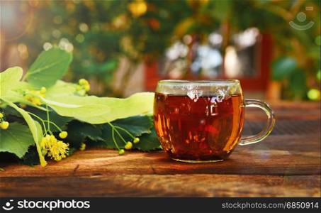 Transparent mug with tea and a lime tree branch on a wooden brown table in the rays of the sun. Transparent mug with tea and a lime tree branch
