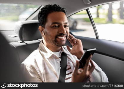 transport, business and technology concept - happy smiling indian male passenger with wireless earphones using smartphone on back seat of taxi car. passenger with earphones and cellphone in taxi car