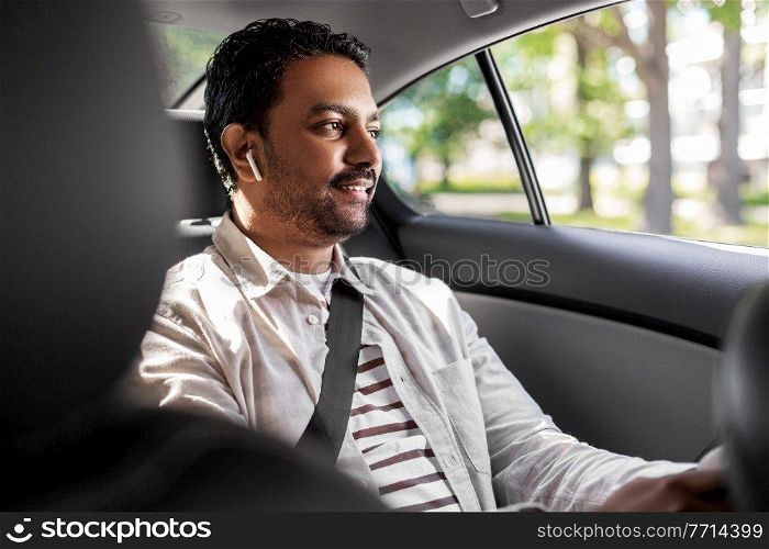 transport, business and technology concept - happy smiling indian male passenger with wireless earphones on back seat of taxi car. indian male passenger with earphones in taxi car