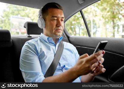 transport, business and technology concept - male passenger or businessman with wireless headphones using smartphone on back seat of taxi car. passenger with headphones using smartphone in car