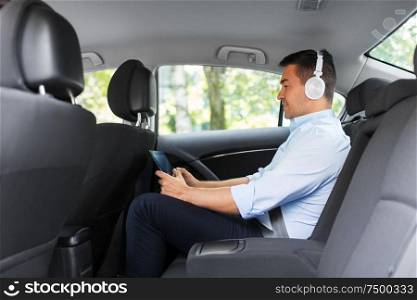 transport, business and technology concept - male passenger or businessman with wireless headphones using tablet pc computer on back seat of taxi car. passenger with headphones using tablet pc in car