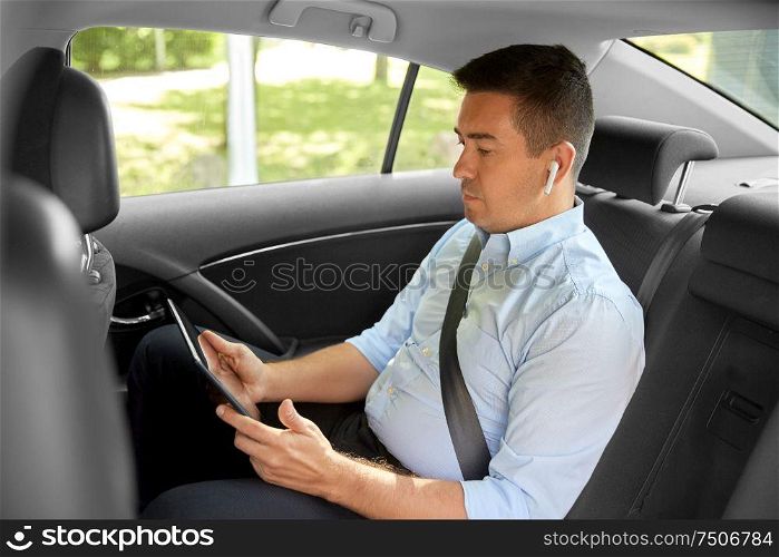 transport, business and technology concept - male passenger or businessman with wireless earphones using tablet pc computer on back seat of taxi car. passenger with earphones and tablet pc in taxi car