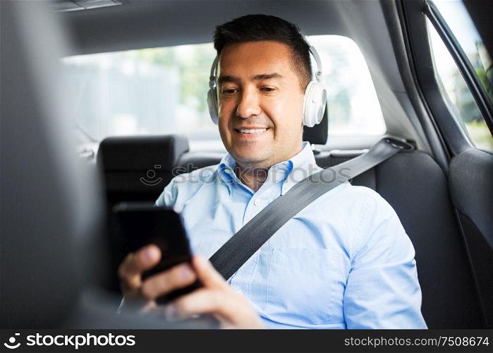 transport, business and technology concept - male passenger or businessman with wireless headphones using smartphone on back seat of taxi car. passenger with headphones using smartphone in car