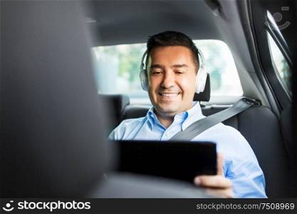 transport, business and technology concept - male passenger or businessman with wireless headphones using tablet pc computer on back seat of taxi car. passenger with headphones using tablet pc in car