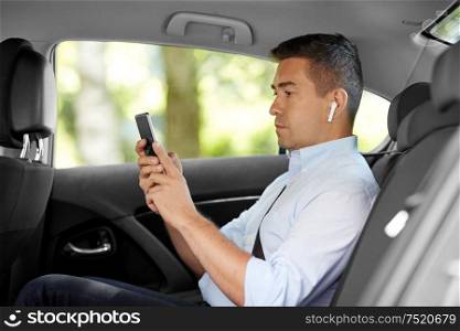 transport, business and technology concept - male passenger or businessman with wireless earphones using smartphone on back seat of taxi car. passenger with earphones and cellphone in taxi car