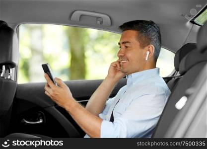 transport, business and technology concept - smiling male passenger or businessman with wireless earphones using smartphone on back seat of taxi car. passenger with earphones and cellphone in taxi car