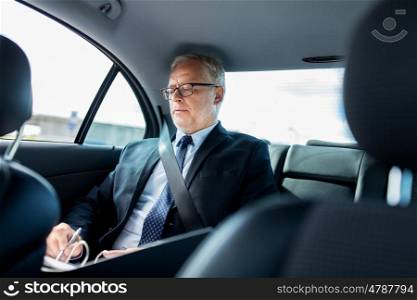 transport, business trip, paperwork and people concept - senior businessman signing papers with pen and driving on car back seat. senior businessman with papers driving in car