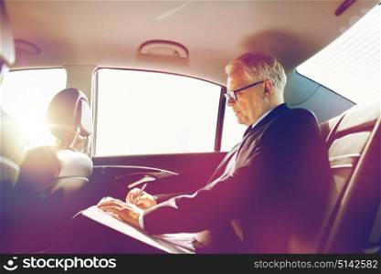 transport, business trip, paperwork and people concept - senior businessman signing papers with pen and driving on car back seat. senior businessman with papers driving in car