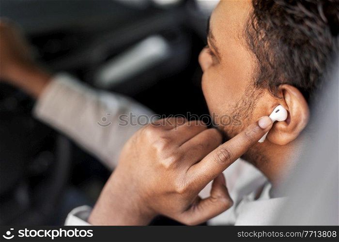 transport, people and technology concept - close up of indian man or driver with wireless earphones or hands free device driving car. man or driver with wireless earphones driving car
