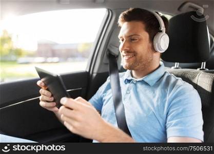 transport, people and technology concept - male passenger with wireless headphones using tablet pc computer on back seat of taxi car. passenger in headphones with tablet pc in taxi car