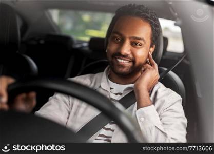 transport, people and technology concept - smiling indian man or driver with wireless earphones or hands free device driving car. man or driver with wireless earphones driving car