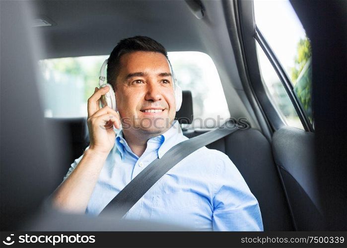 transport, technology and people concept - male passenger or businessman with wireless headphones listening to music on back seat of taxi car. passenger in headphones listening to music in car