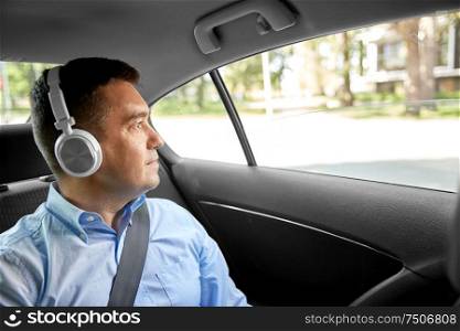 transport, technology and people concept - male passenger or businessman with wireless headphones listening to music on back seat of taxi car. passenger in headphones listening to music in car
