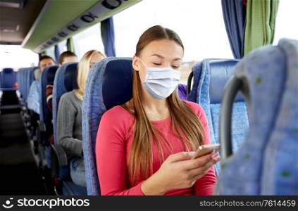transport, tourism, road trip and people concept - young woman wearing face protective medical mask for protection from virus disease sitting in travel bus with smartphone. woman in mask with smartphone in travel bus