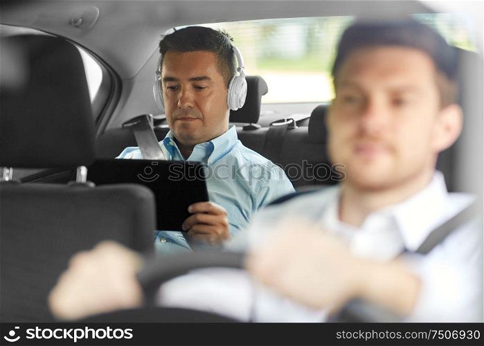 transportation, taxi and technology concept - middle aged male passenger with tablet pc computer and headphones on back seat and car driver. man with tablet pc and headphones riding in car