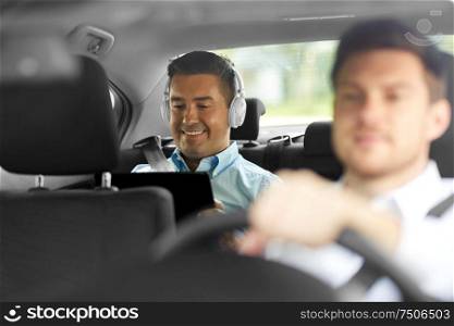 transportation, taxi and technology concept - smiling middle aged male passenger with tablet pc computer and headphones on back seat and car driver. man with tablet pc and headphones riding in car
