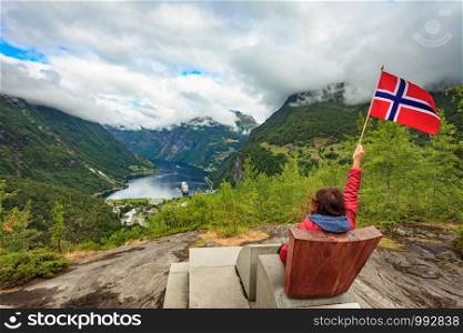 Travel adventure concept. Female tourist enjoying view over Geirangerfjord from Flydalsjuvet viewpoint, holding norwegian flag.. Tourist over Geiranger fjord holds norwegian flag