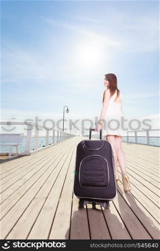Travel, adventure, packing. Young woman wearing short white dress walking with suitcase on wheels through wooden pier. Young woman walking with suitcase on wheels