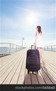Travel, adventure, packing. Young woman wearing short white dress walking with suitcase on wheels through wooden pier. Young woman walking with suitcase on wheels