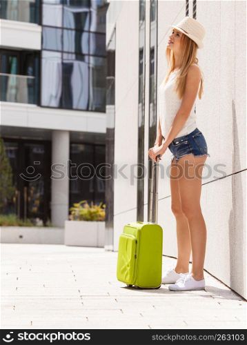 Travel, adventure, teenage journey concept. Walking woman wearing denim shorts, white top and sun hat suitcase holding suitcase on wheels. Attractive woman with suitcase walking after arrival