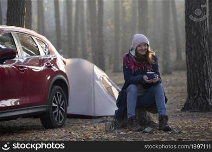 travel by car. girl tourist sits on a tree stump with a cup of tea and a car with a tent in the background