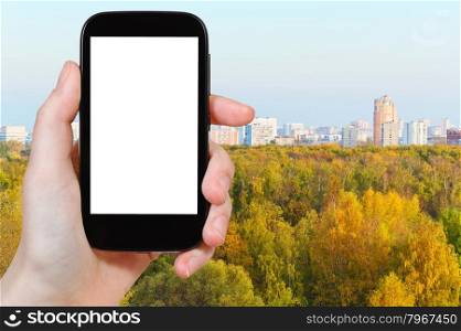travel concept - hand holds smartphone with cut out screen and autumn forest and city on background