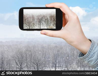 travel concept - tourist takes picture of oak and birch trees in snow blizzard in forest in spring on smartphone