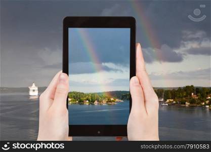 travel concept - tourist takes picture of rainbow over small village on Baltic sea coast on smartphone, Sweden