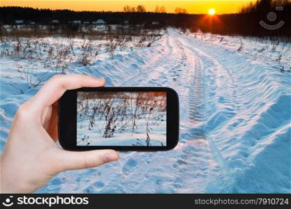travel concept - tourist takes picture of sunset over blue winter snowdrifts on country field on smartphone,