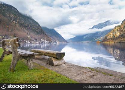 Travel destinations theme image with the Dachstein Mountains reflected in the water of the Hallstatter lake and wooden benches on its shore, in Hallstatt, Austria.