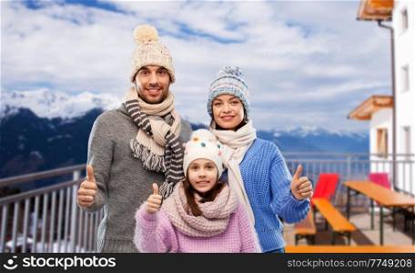 travel, family and winter holidays concept - happy mother, father and little daughter in knitted hats and scarves showing thumbs up over mountains and ski resort background. happy family showing thumbs up in winter