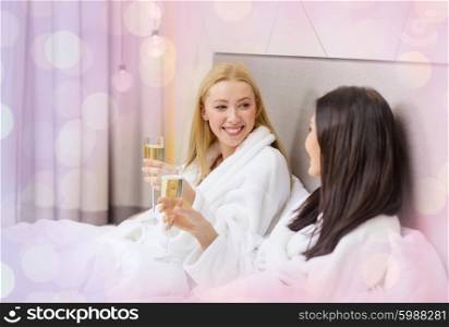 travel, friendship, bachelorette party and people concept - smiling girlfriends in bathrobes with champagne glasses in bed over holidays lights background