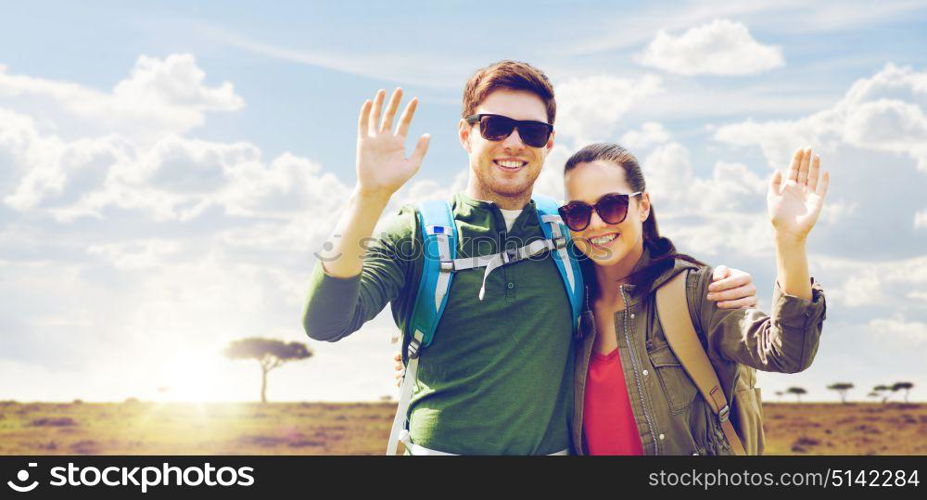 travel, hiking, backpacking, tourism and people concept - happy couple with backpacks waving hands over african savannah background. smiling couple with backpacks traveling in africa