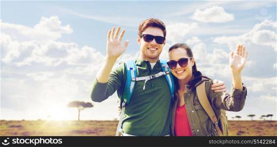 travel, hiking, backpacking, tourism and people concept - happy couple with backpacks waving hands over african savannah background. smiling couple with backpacks traveling in africa