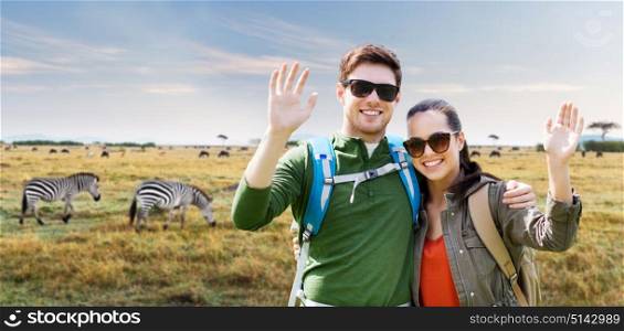 travel, hiking, backpacking, tourism and people concept - happy couple with backpacks waving hands over african savannah and zebras background. smiling couple with backpacks traveling in africa