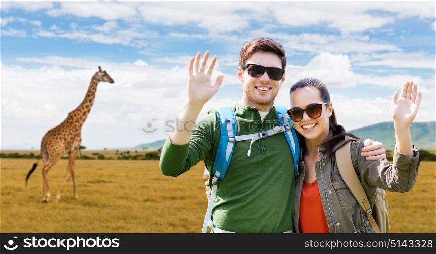 travel, hiking, backpacking, tourism and people concept - happy couple with backpacks waving hands over african savannah and giraffe background. smiling couple with backpacks traveling in africa