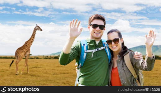 travel, hiking, backpacking, tourism and people concept - happy couple with backpacks waving hands over african savannah and giraffe background. smiling couple with backpacks traveling in africa