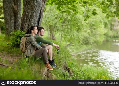 travel, hiking, backpacking, tourism and people concept - smiling couple with backpacks resting on river bank in nature on river bank