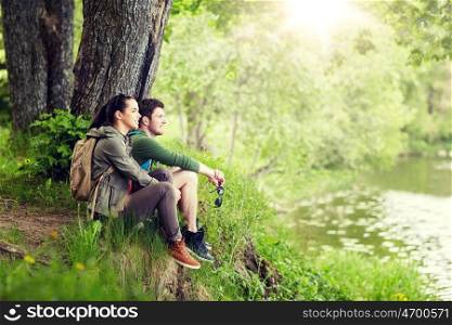 travel, hiking, backpacking, tourism and people concept - smiling couple with backpacks resting on river bank in nature on river bank. smiling couple with backpacks in nature. smiling couple with backpacks in nature