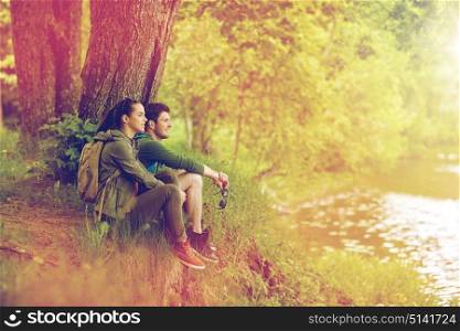 travel, hiking, backpacking, tourism and people concept - smiling couple with backpacks resting on river bank in nature on river bank. smiling couple with backpacks in nature