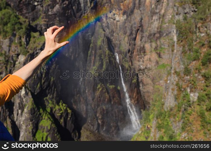 Travel, holidays. Tourist woman at Voringsfossen waterfall. holding rainbow in hand. Mabodalen canyon Norway. National tourist Hardangervidda route, Eidfjord sightseeing tour.. Tourist woman by Voringsfossen waterfall, Norway
