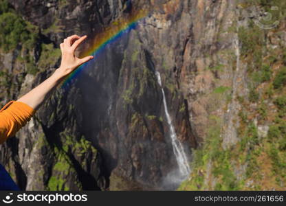 Travel, holidays. Tourist woman at Voringsfossen waterfall. holding rainbow in hand. Mabodalen canyon Norway. National tourist Hardangervidda route, Eidfjord sightseeing tour.. Tourist woman by Voringsfossen waterfall, Norway