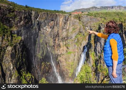 Travel, holidays. Tourist woman looking at Voringsfossen waterfall. Mabodalen canyon Norway. National tourist Hardangervidda route, Eidfjord sightseeing tour.. Tourist woman by Voringsfossen waterfall, Norway