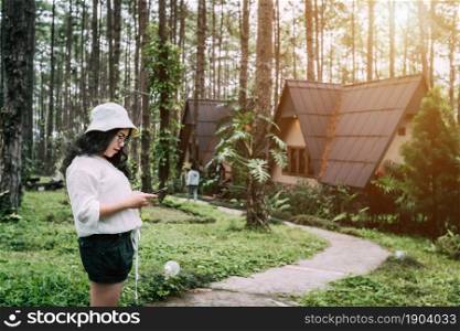 travel relax vacation,Asian female tourist in white dress with hat stands writing smartphone in pinewood cabin in pine forest green on nature trail at Doi Bo Luang Forest Park,Chiang Mai, Thailand