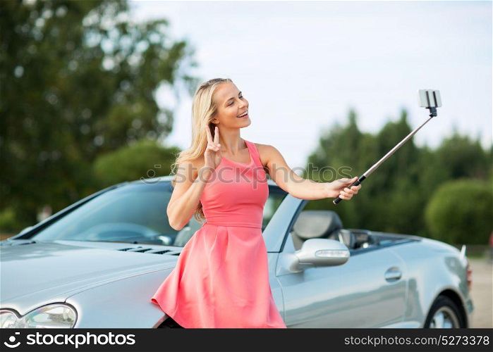 travel, road trip and people concept - happy young woman at convertible car taking picture by smartphone selfie stick and showing peace hand sign. woman taking picture by selfie stick at car