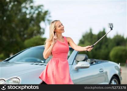 travel, road trip and people concept - happy young woman at convertible car taking picture by smartphone selfie stick and showing peace hand sign. woman taking picture by selfie stick at car